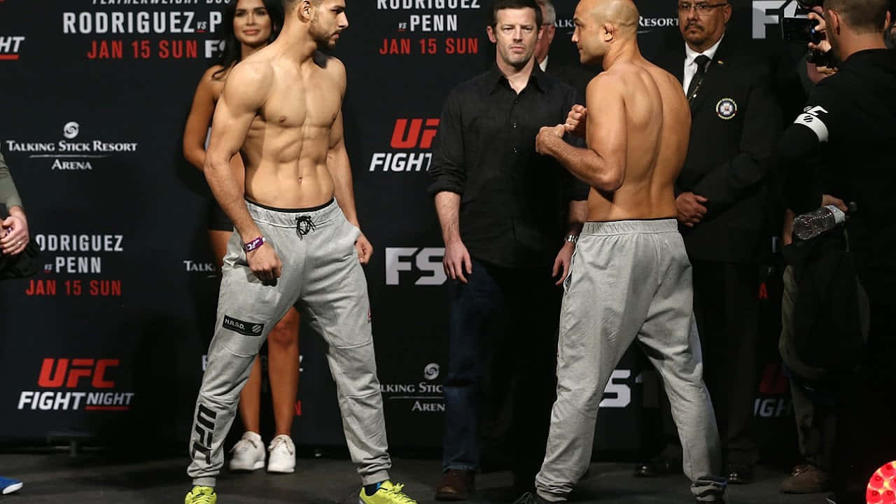 UFC 桌布 壁紙 — 兩位肌肉發達的UFC拳手在明亮的舞台燈光下，於稱重活動中對峙，身後有活動品牌標誌。