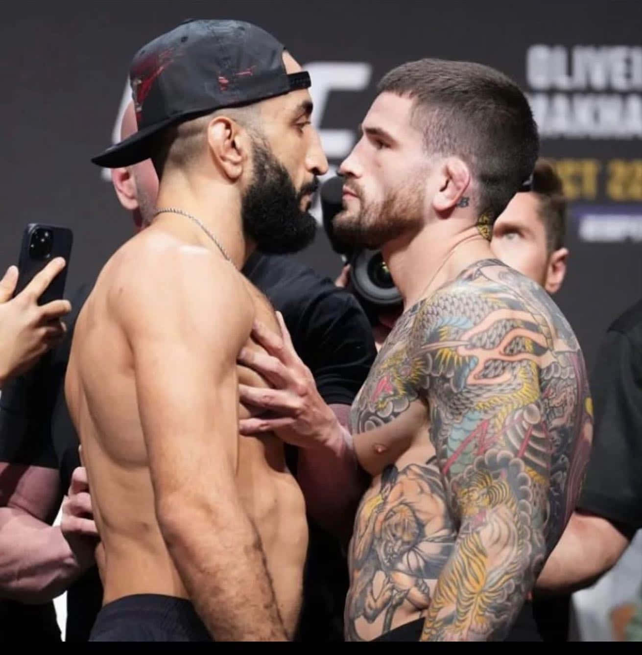 MMA 桌布 壁紙 — 兩位肌肉發達的UFC拳手在稱重時激烈對峙，身上有刺青，戴著帽子，在明亮的場館燈光下。