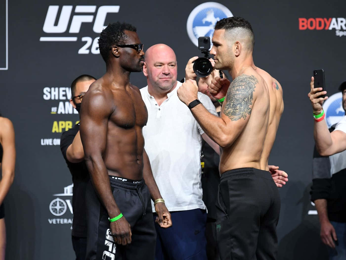 MMA 桌布 壁紙 — 兩位肌肉發達的UFC拳手在稱重活動中對峙，相機閃光燈閃爍，工作人員在旁。