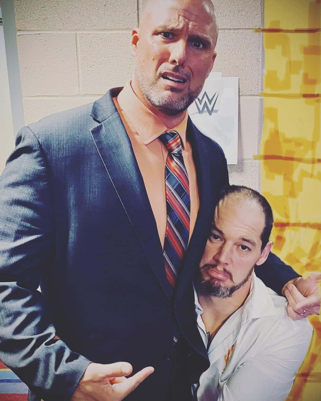 WWE 桌布 壁紙 — 兩位肌肉發達的WWE摔角手在後台走廊緊密擺姿勢，一位穿西裝，另一位穿白襯衫，背後可見WWE標誌。