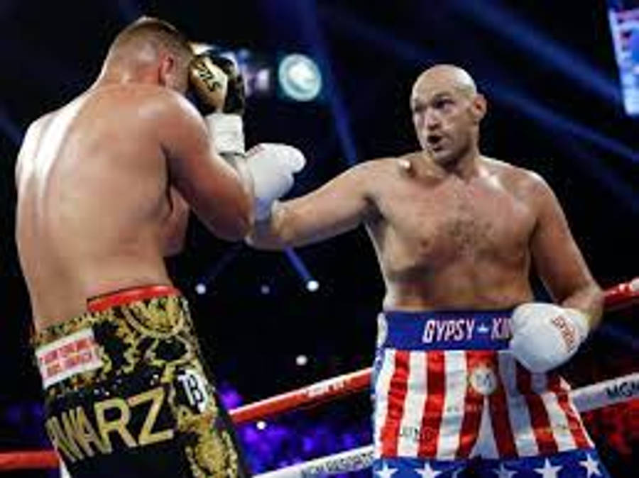 Tyson Fury 桌布 壁紙 — 兩位肌肉發達的拳擊手在燈光明亮的擂台上交鋒，一人穿金色短褲，另一人穿紅白藍星紋短褲。