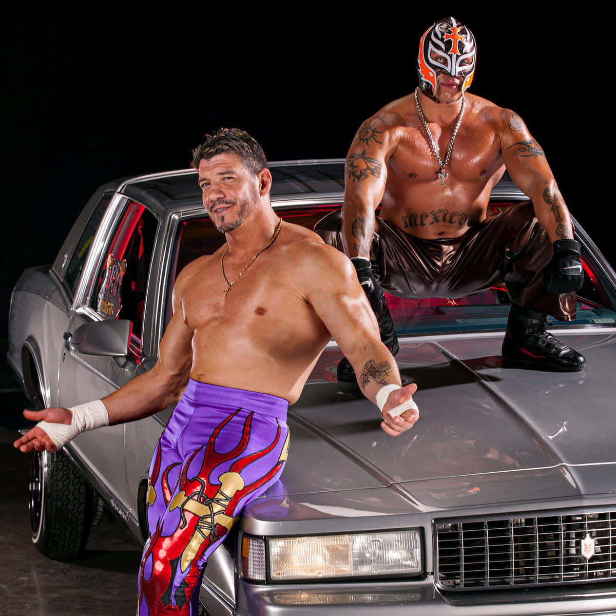 Wrestling 桌布 壁紙 — 兩位肌肉發達的摔角手，一位穿著紫色火焰緊身褲，另一位戴著面具，在黑色背景前自信地擺姿勢，旁邊是一輛銀色汽車。