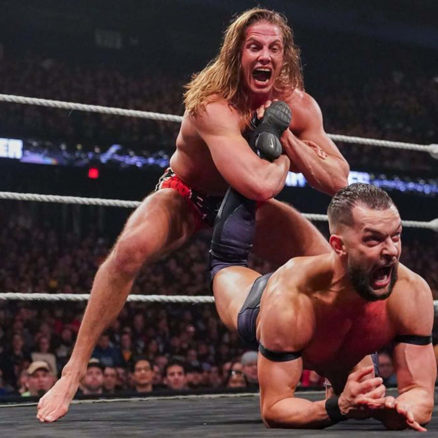 WWE 桌布 壁紙 — 兩位肌肉發達的摔角手在燈光明亮的擂台上激烈糾纏，背景是喧囂的人群。