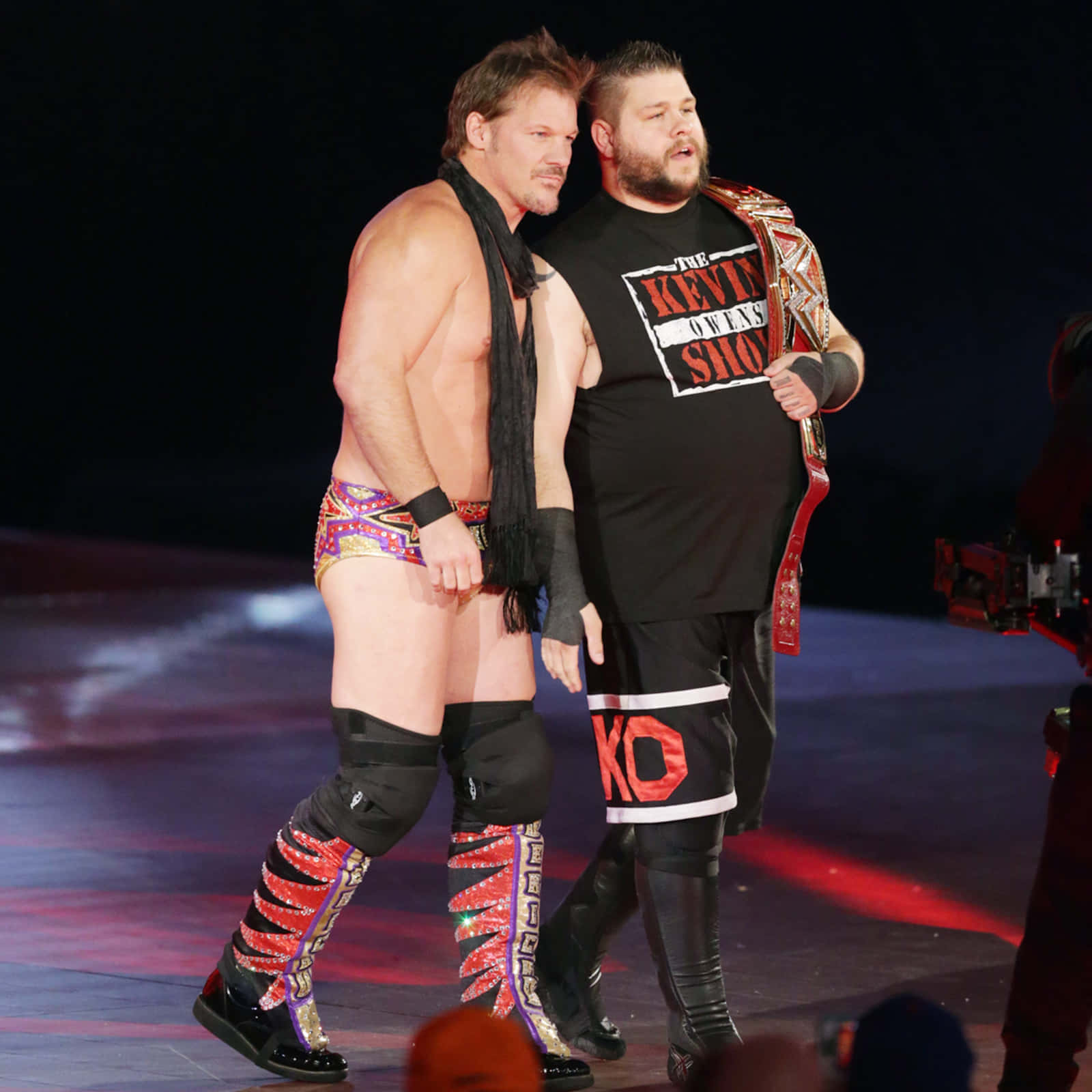 WWE 桌布 壁紙 — 兩位肌肉發達的摔角手在燈光璀璨的舞台上並肩而行，一人赤裸上身，穿著色彩繽紛的靴子，另一人穿著黑色T恤，手持冠軍腰帶。