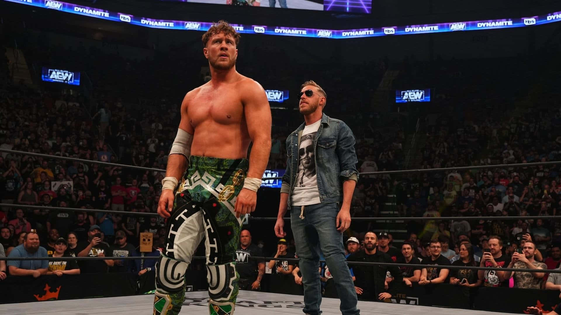 Aew 桌布 壁紙 — 兩位肌肉發達的摔角手站在燈光明亮的擂台上，周圍是AEW Dynamite賽事中喧鬧的人群。