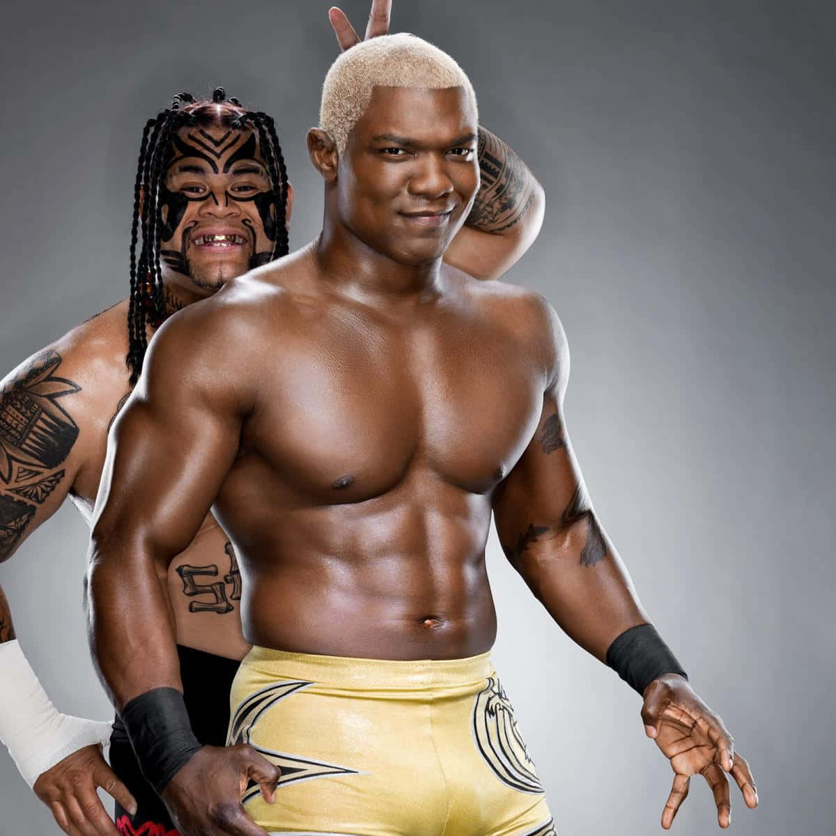 WWE 桌布 壁紙 — 兩位肌肉發達的摔角手自信地擺姿勢，背景是灰色攝影棚，一位金髮穿黃色短褲，另一位有部落彩繪和辮子。