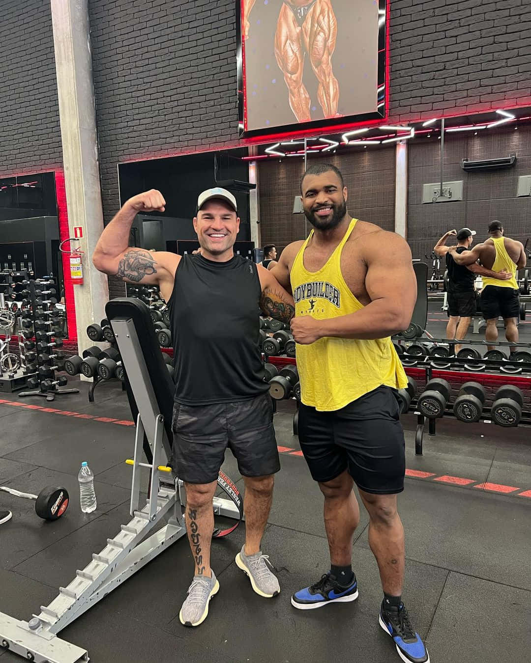 Champion 桌布 壁紙 — 兩位肌肉發達的男子在現代健身房中自豪地展示肌肉，背景有啞鈴和健美運動員海報。