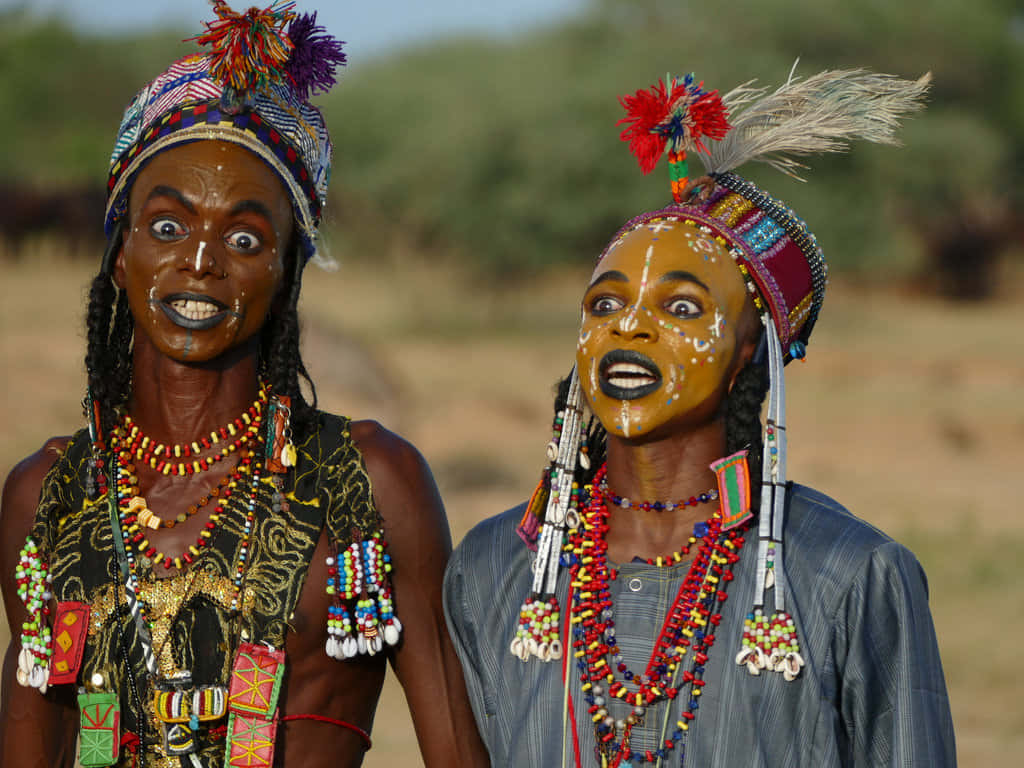 African Culture 桌布 壁紙 — 兩位臉上塗有顏料、佩戴色彩繽紛珠飾的女性，在明亮的日光下站在乾燥的草原上。