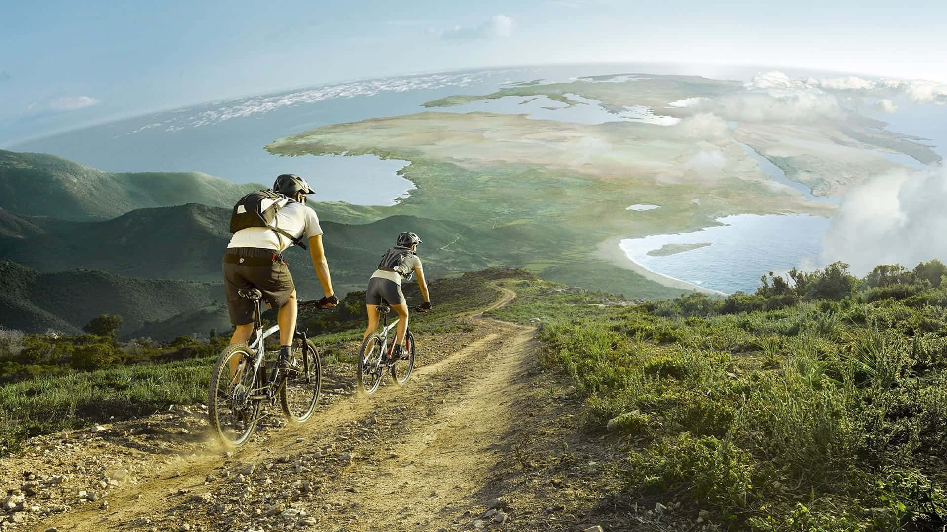 Bikes 桌布 壁紙 — 兩位自行車手在泥土路上踩踏爬坡，俯瞰廣闊的綠色山谷和海岸水域，天空湛藍明亮。