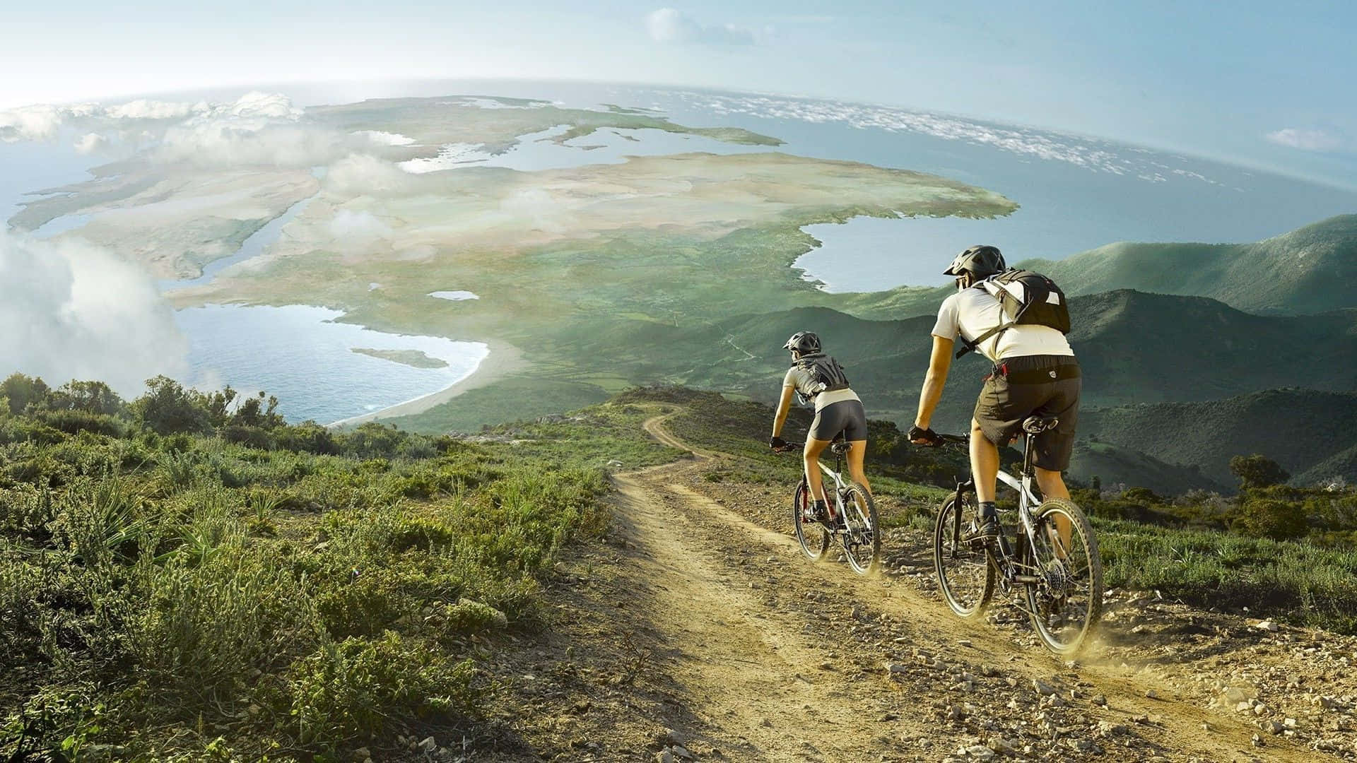 Mtb 桌布 壁紙 — 兩位自行車騎士在泥土路上踩踏上山，俯瞰廣闊的沿海景觀，蜿蜒的水道和綠色的山丘在明亮的天空下。