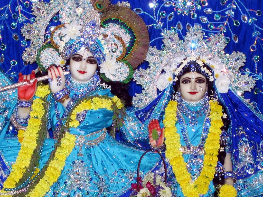 Hindu God 桌布 壁紙 — 兩位裝飾華麗的印度教神祇，身穿閃爍藍色服飾和鑲有寶石的皇冠，優雅地坐在深藍色珠飾背景前。