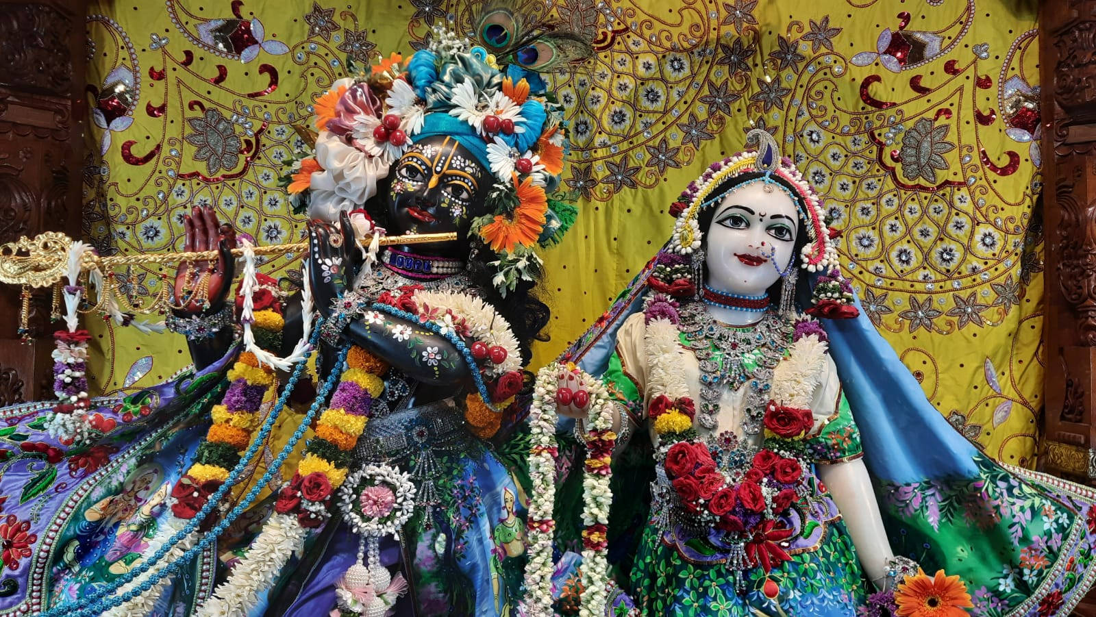Iskcon 桌布 壁紙 — 兩位裝飾華麗的印度教神祇並排坐在鮮豔的黃色圖案背景前，披著色彩繽紛的布料和花環。