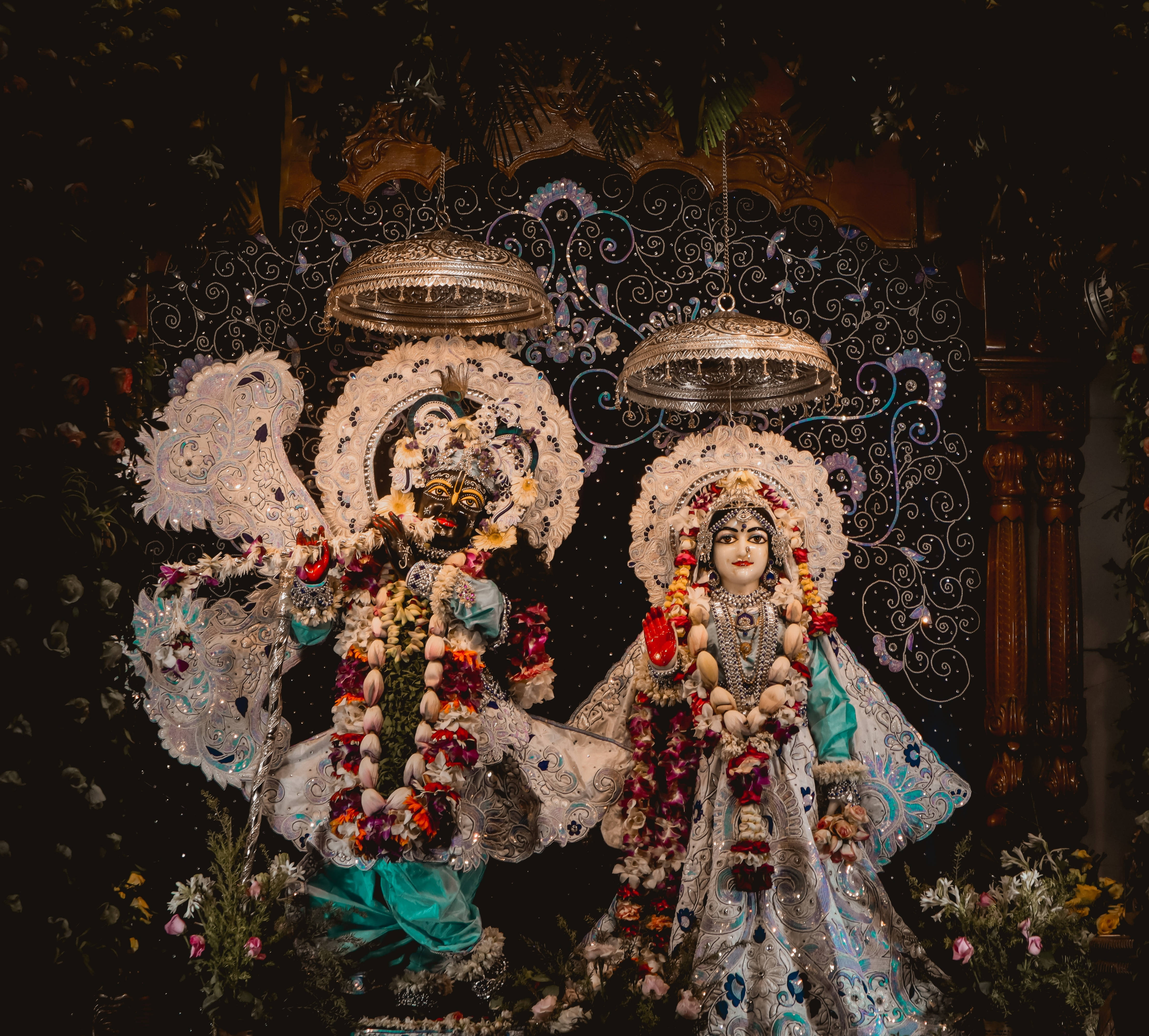 Hindu Temple 桌布 壁紙 — 兩位裝飾華麗的印度教神靈坐在裝飾華麗的華蓋下，置身於昏暗寺廟環境中的花卉裝飾之中。