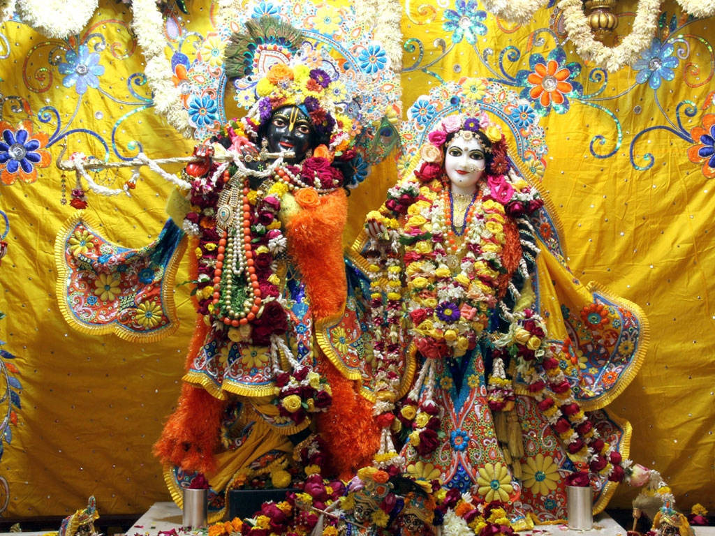 Iskcon 桌布 壁紙 — 兩位裝飾華麗的印度教神靈站在充滿活力的黃色花卉背景前，披著色彩繽紛的花環和華麗的服飾。