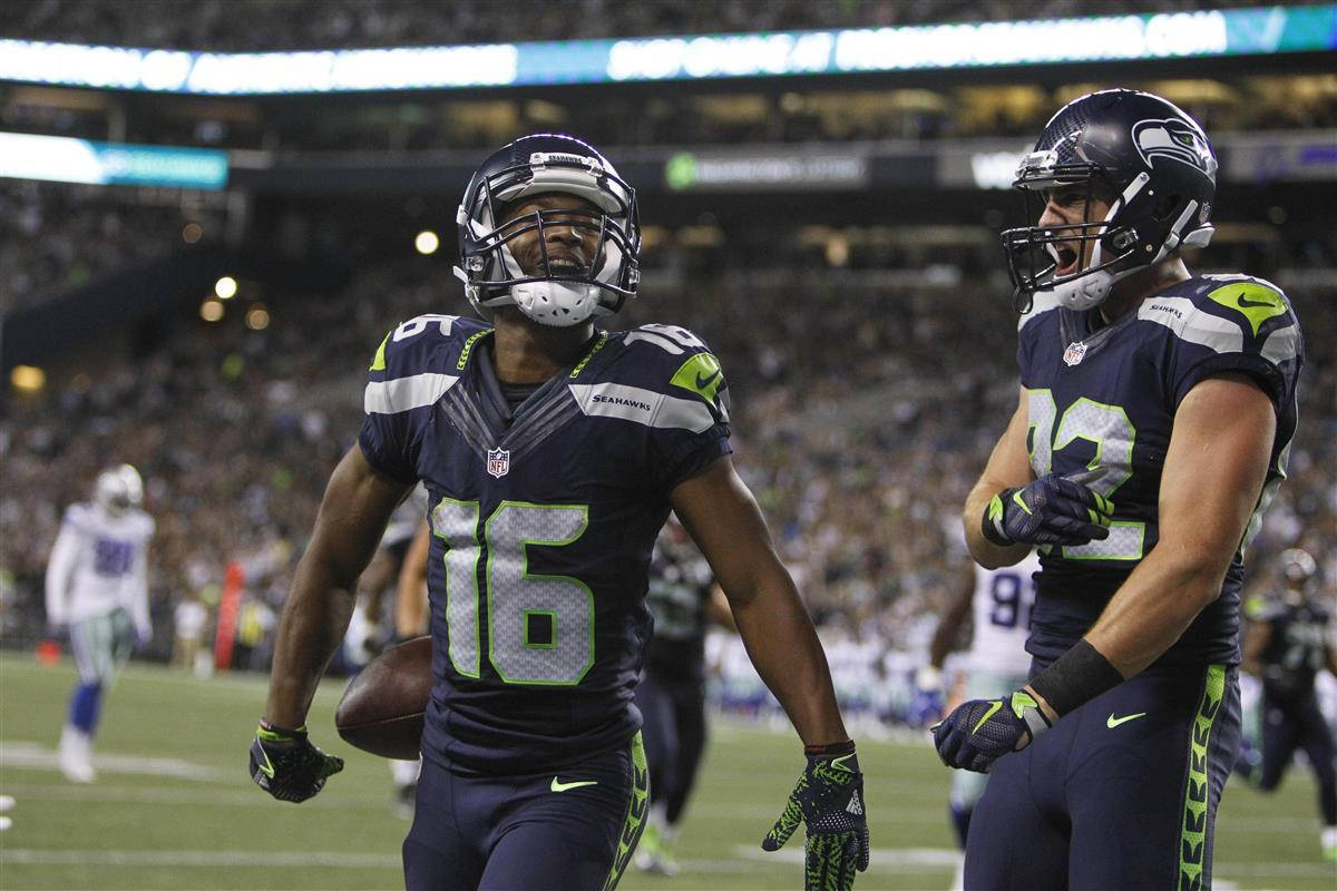 Seattle Seahawks 桌布 壁紙 — 兩位西雅圖海鷹隊美式足球球員在燈光明亮的球場上慶祝達陣，背景有歡呼的球迷。
