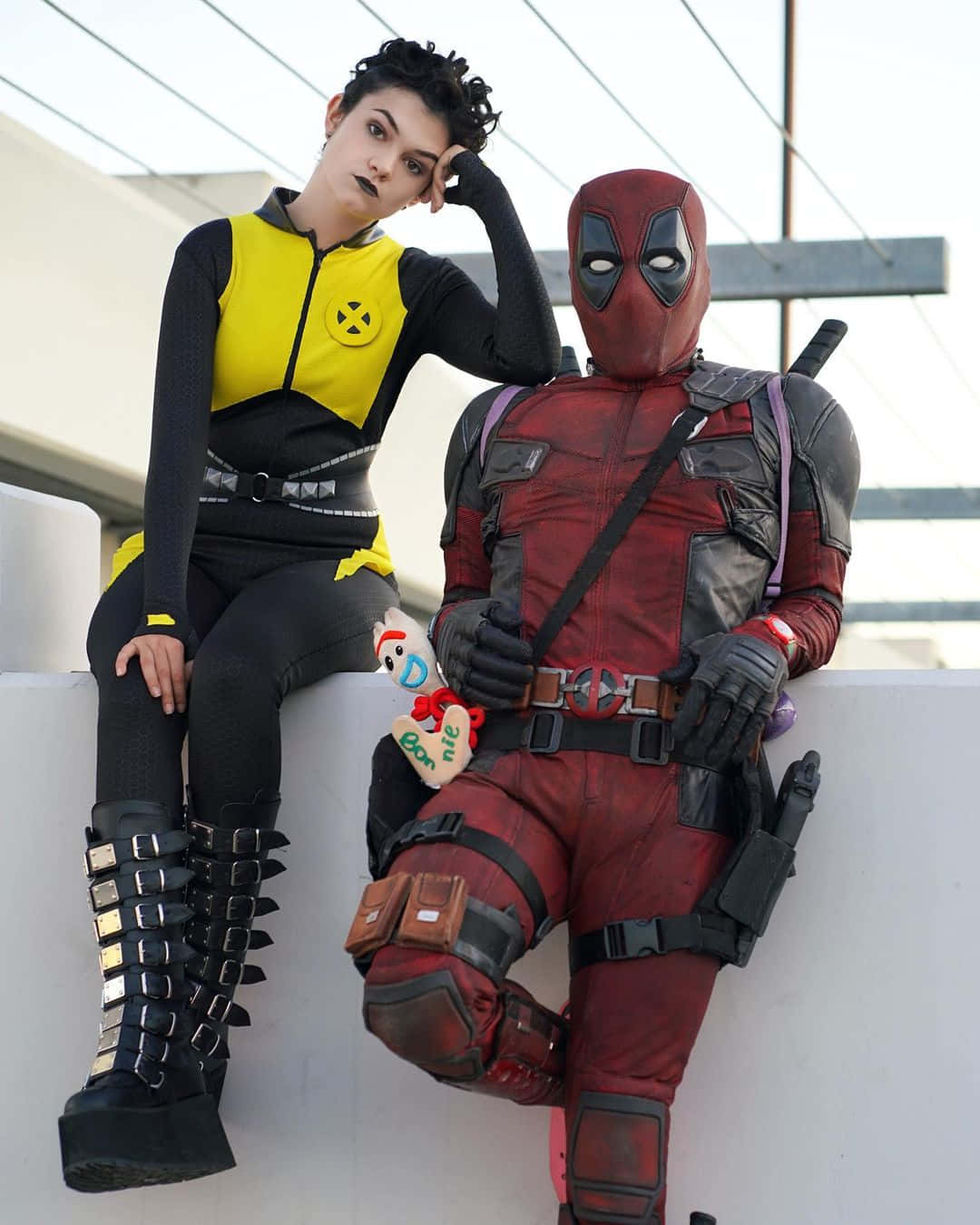 Deadpool And Negasonic Teenage Warhead 桌布 壁紙 — 兩位角色扮演者，一位扮演Jean Grey，一位扮演Deadpool，在明亮的戶外背景下隨意地坐在白色邊緣上擺姿勢。