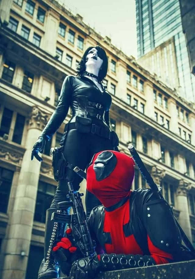 Deadpool And Domino 桌布 壁紙 — 兩位角色扮演者，一位身穿黑色皮革站立，另一位身穿紅黑色蹲下並手持武器，身處城市街道。