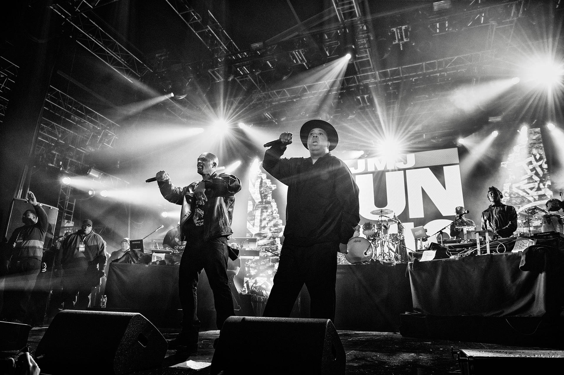 band 桌布 壁紙 — 兩位說唱歌手在燈光昏暗的舞台上，沐浴在戲劇性的聚光燈下，身後有樂隊，充滿活力地表演。