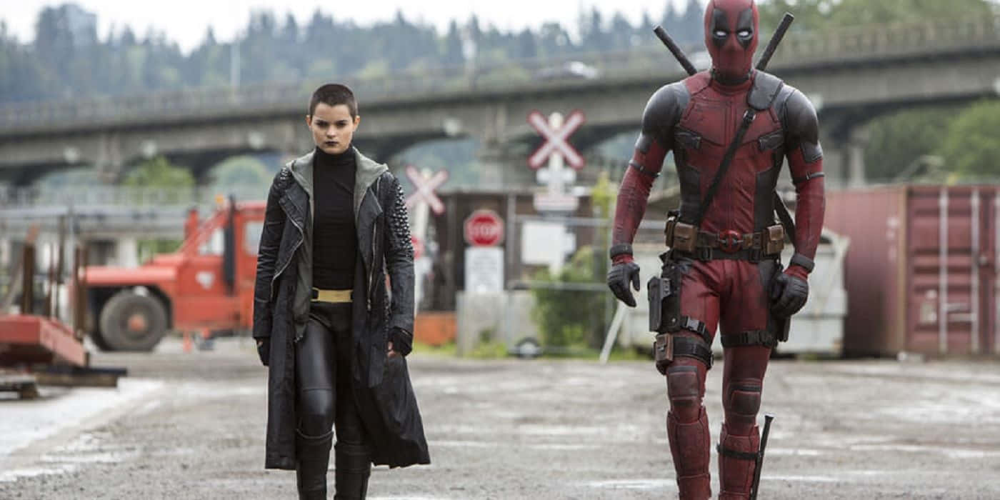 Deadpool And Negasonic Teenage Warhead 桌布 壁紙 — 兩位超級英雄死侍和多米諾站在一個灰暗的工業場地，天空多雲，背後有橋樑和火車軌道。