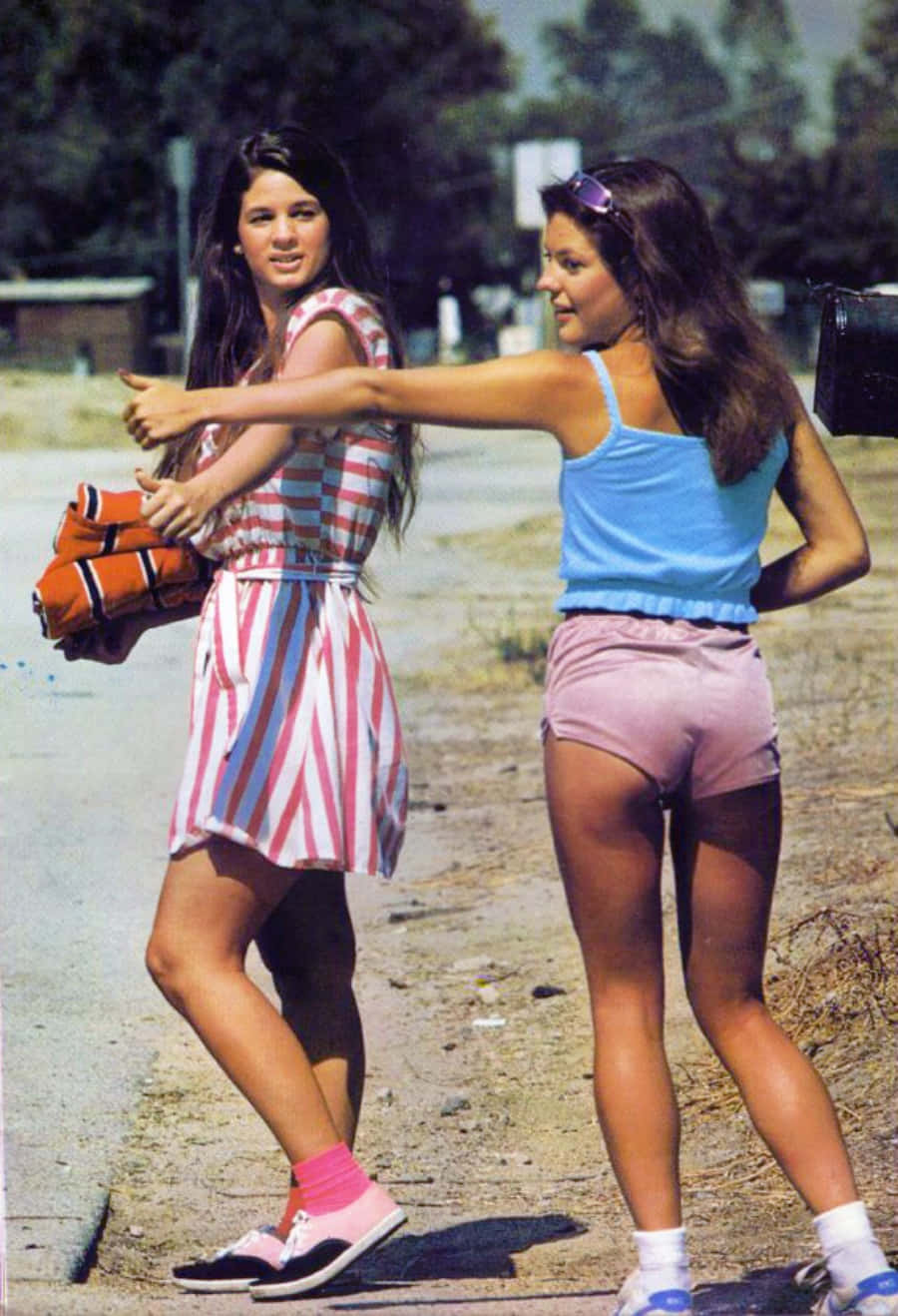 70年代動圖 桌布 壁紙 — 兩位身穿1970年代復古服裝的年輕女性在陽光明媚的路邊，以樹木和遠處的建築物為背景，俏皮地擺姿勢。