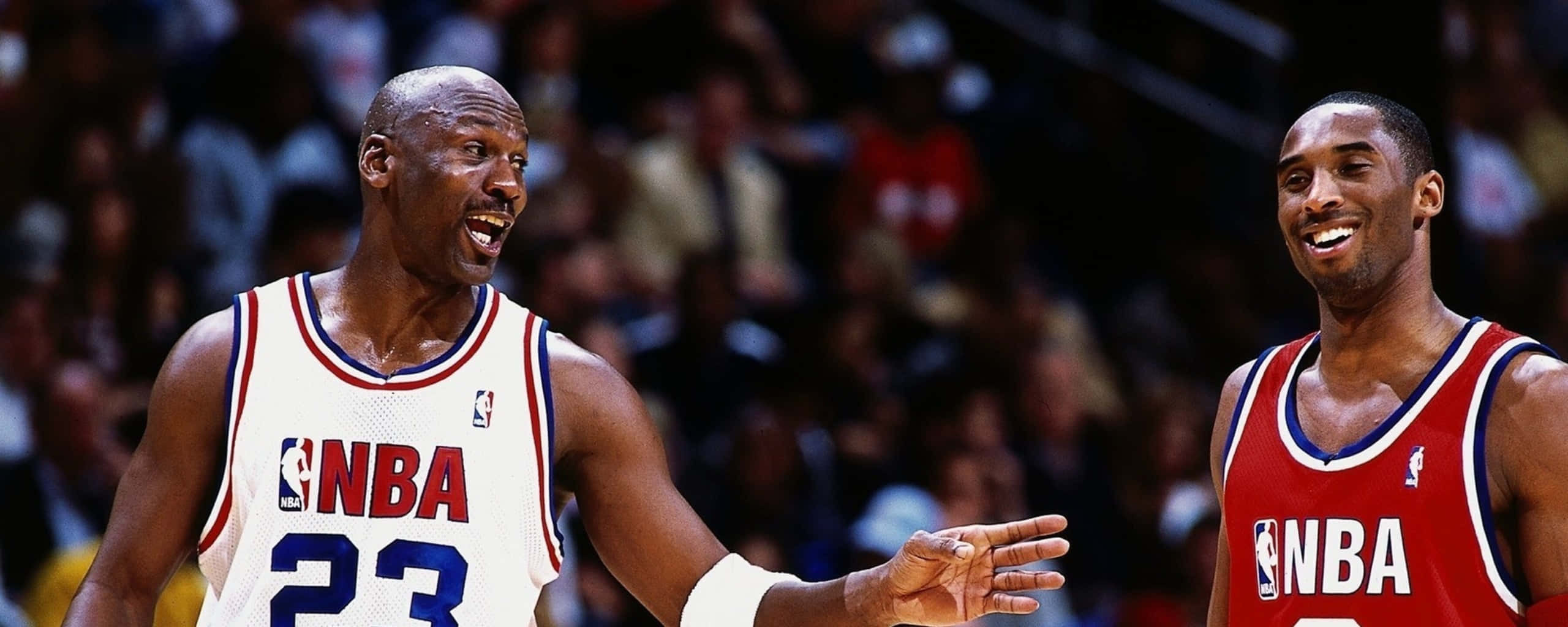 Jordan And Kobe 桌布 壁紙 — 兩位身穿NBA球衣的傳奇籃球運動員在比賽中分享歡樂時刻，背景是模糊的人群。
