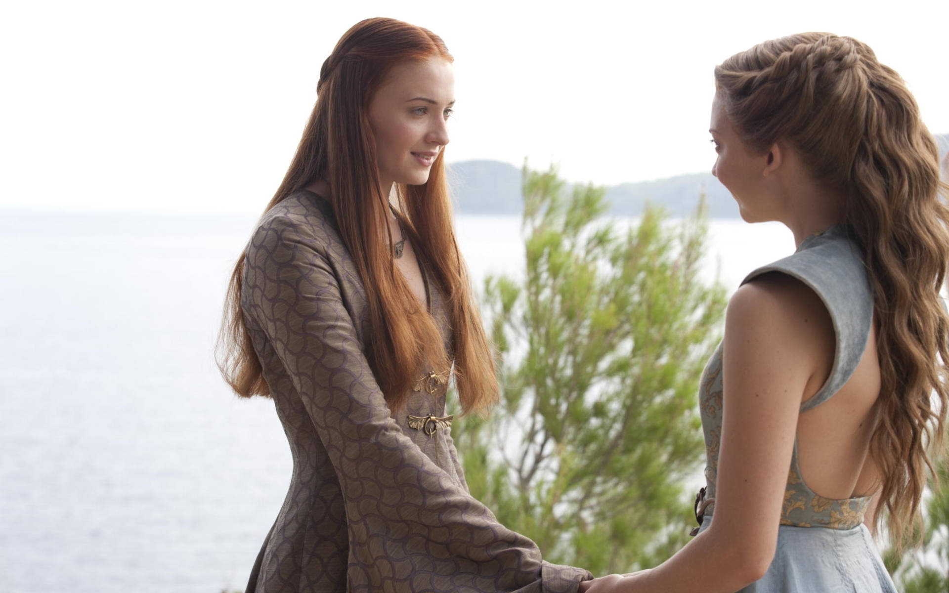 Sansa Stark 桌布 壁紙 — 兩位身穿中世紀風格連衣裙的女性手牽手，在綠意盎然和水邊的海岸背景下相視而笑。