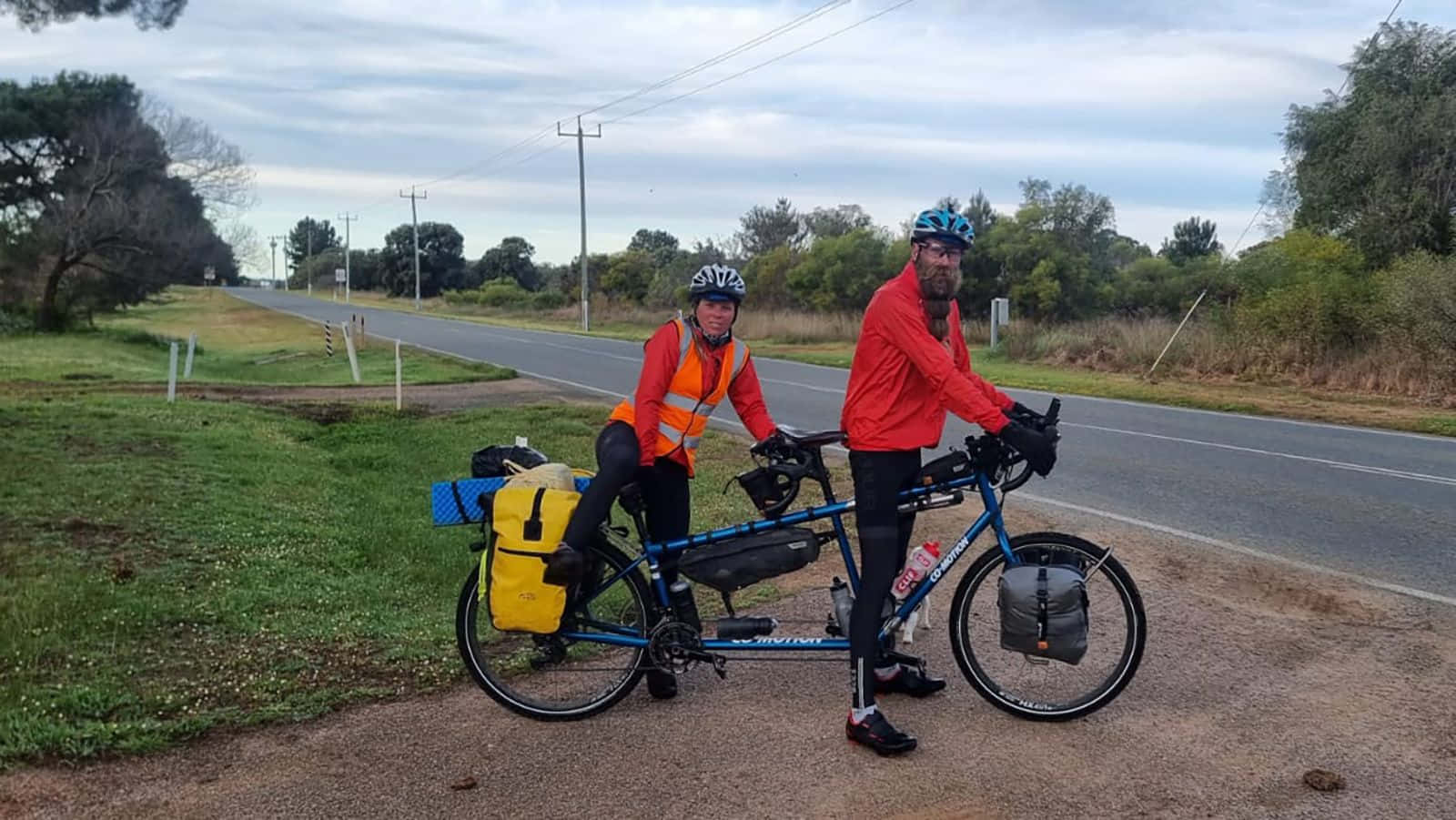 bike riding 桌布 壁紙 — 兩位身穿亮紅色夾克的自行車騎士在陰天時停在鄉間道路上，旁邊是一輛載滿裝備的雙人自行車。