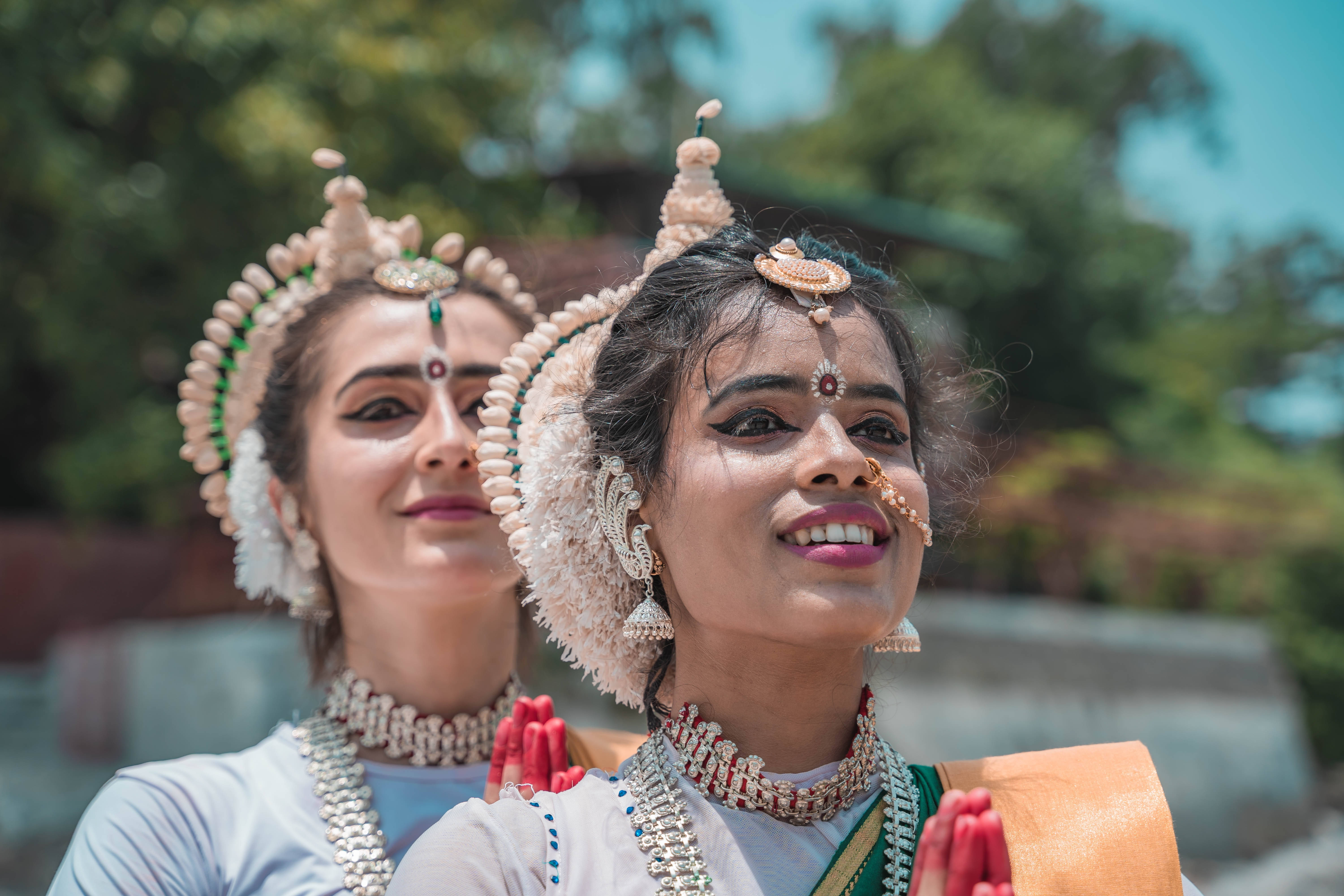 ganesh 4K 桌布 壁紙 — 兩位身穿傳統印度舞蹈服飾、佩戴華麗頭飾和珠寶的女性，在陽光普照的綠色背景下優雅地微笑。