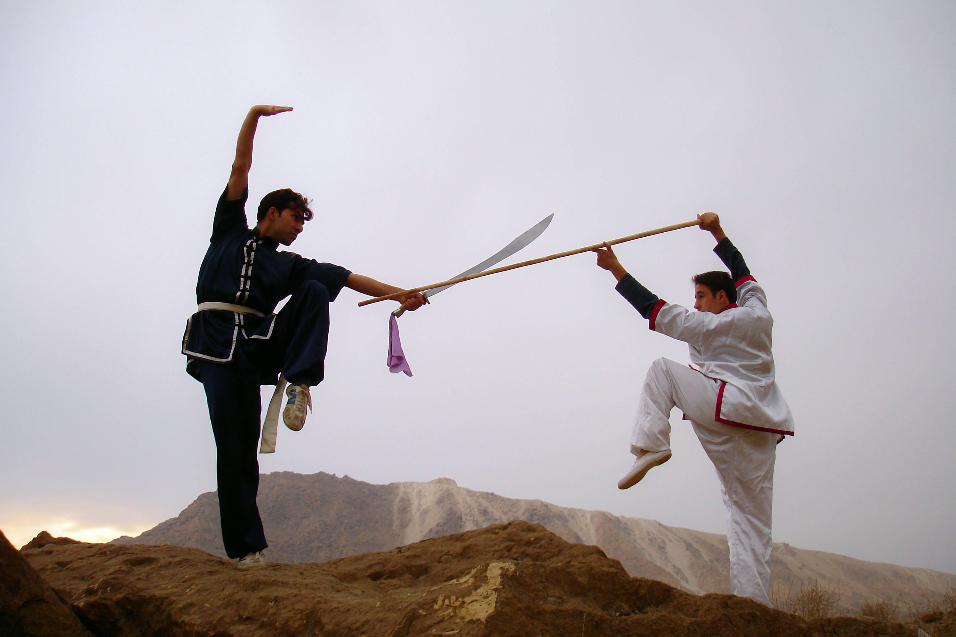 Exercise 桌布 壁紙 — 兩位身穿傳統服裝的武術家在黃昏時分的岩石山坡上以劍和棍對決。