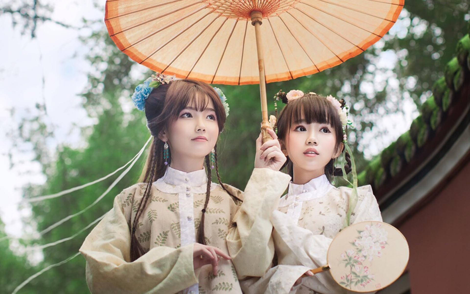 模特兒 桌布 壁紙 — 兩位身穿傳統漢服的年輕女性在明亮的天空下拿著一把大橙色遮陽傘和一把花卉扇子。