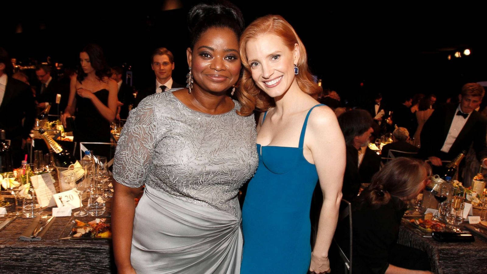 jessica chastain 桌布 壁紙 — 兩位身穿優雅晚禮服的女士在燭光餐桌和背景賓客的正式晚宴上溫暖微笑。