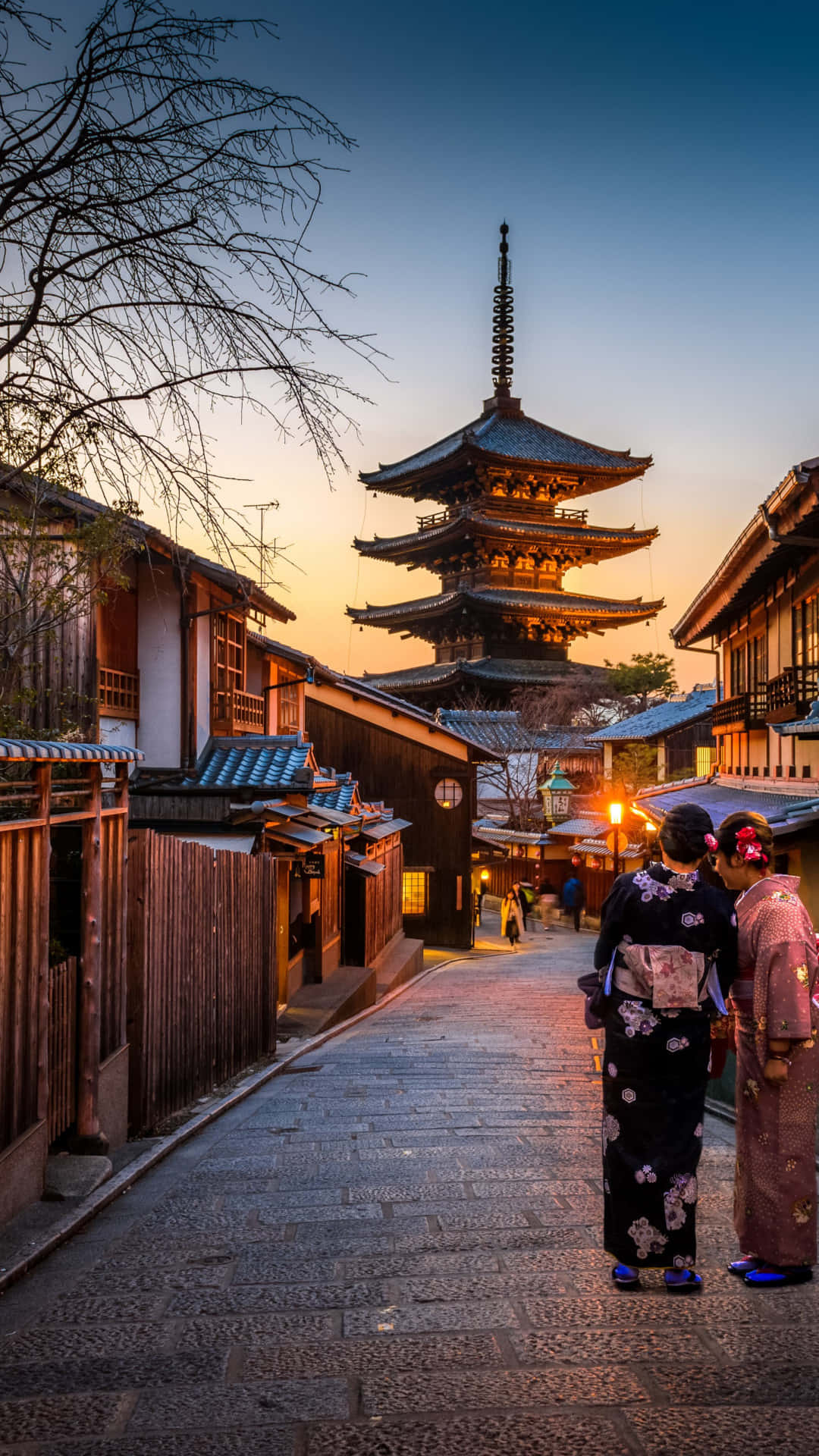 日本 桌布 壁紙 — 兩位身穿和服的女士沿著石徑漫步，走向高聳的寶塔，金色的夕陽灑滿傳統的日本街道。