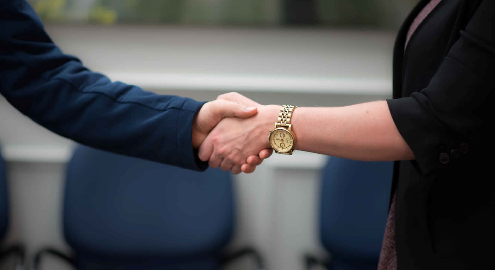 hands 桌布 壁紙 — 兩位身穿商務服裝的專業人士握手，其中一隻手腕上可見一隻金錶，背景是模糊的辦公室。
