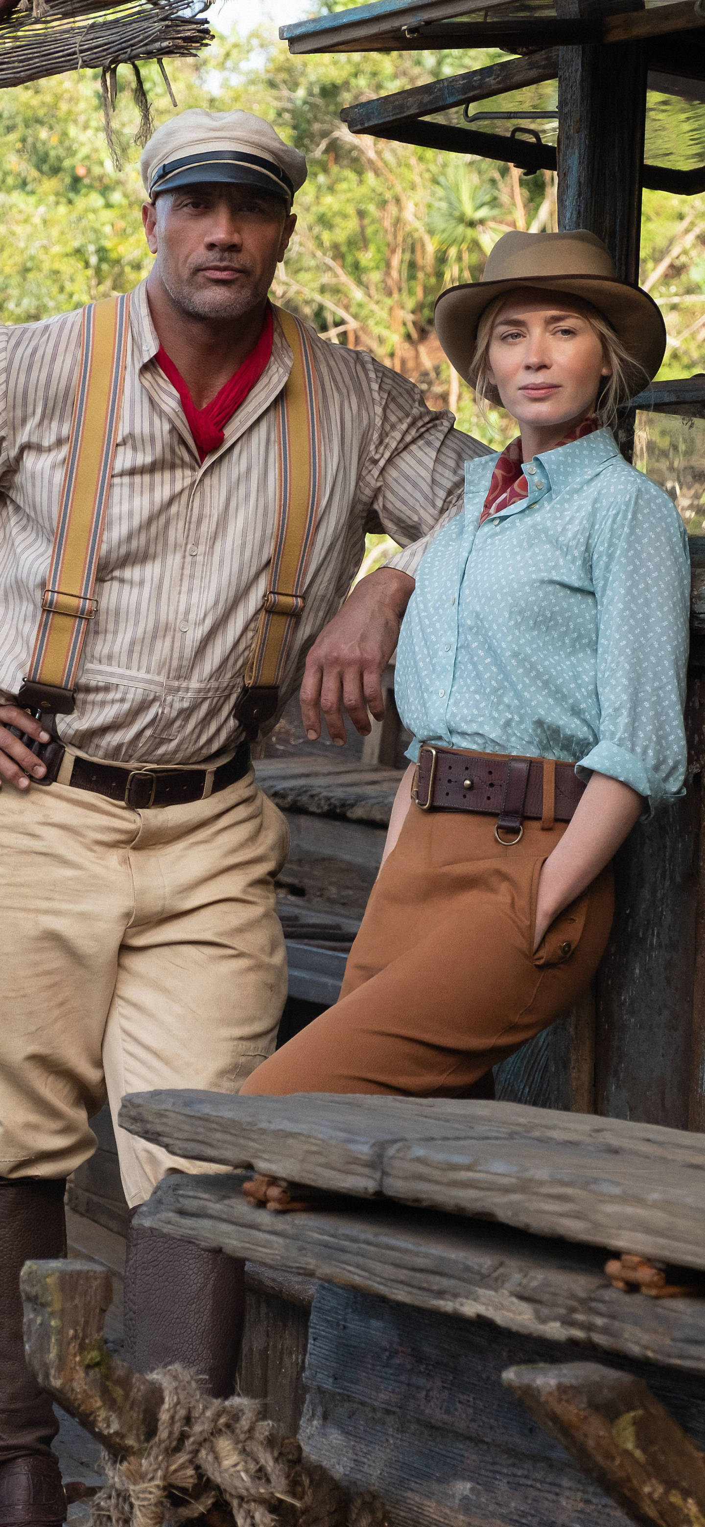 Jungle Cruise 桌布 壁紙 — 兩位身穿復古探險家服裝的演員自信地站在茂密叢林中的一座質樸木結構旁。