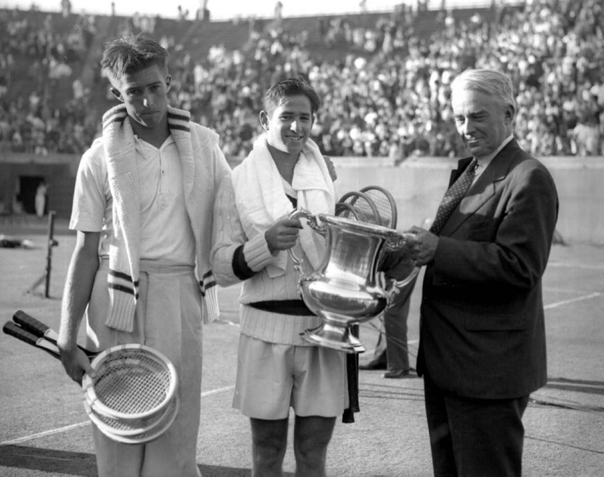 Tennis 桌布 壁紙 — 兩位身穿復古服裝的年輕網球選手站在陽光普照的球場上，在擠滿觀眾的人群前，由一名官員向他們頒發獎盃。