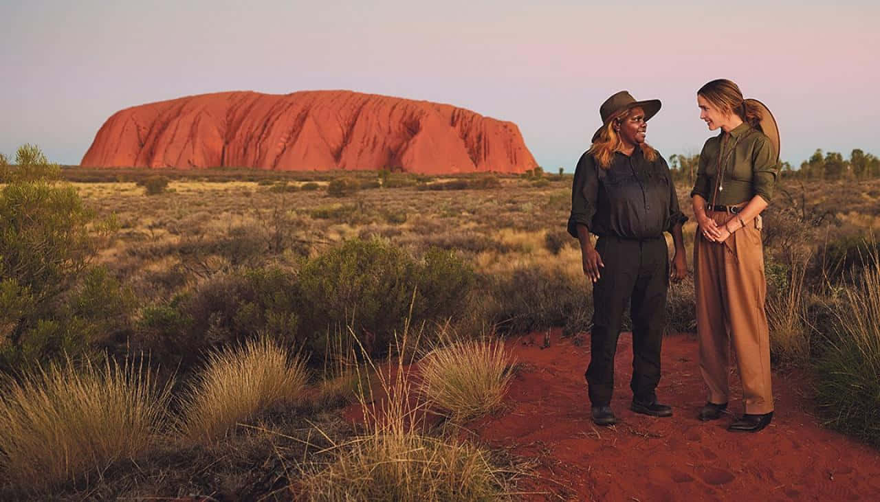 Aboriginal 桌布 壁紙 — 兩位身穿探險家服飾的女性在日落時分，於巨大的紅色單體岩烏魯魯前的紅色泥土小徑上交談。