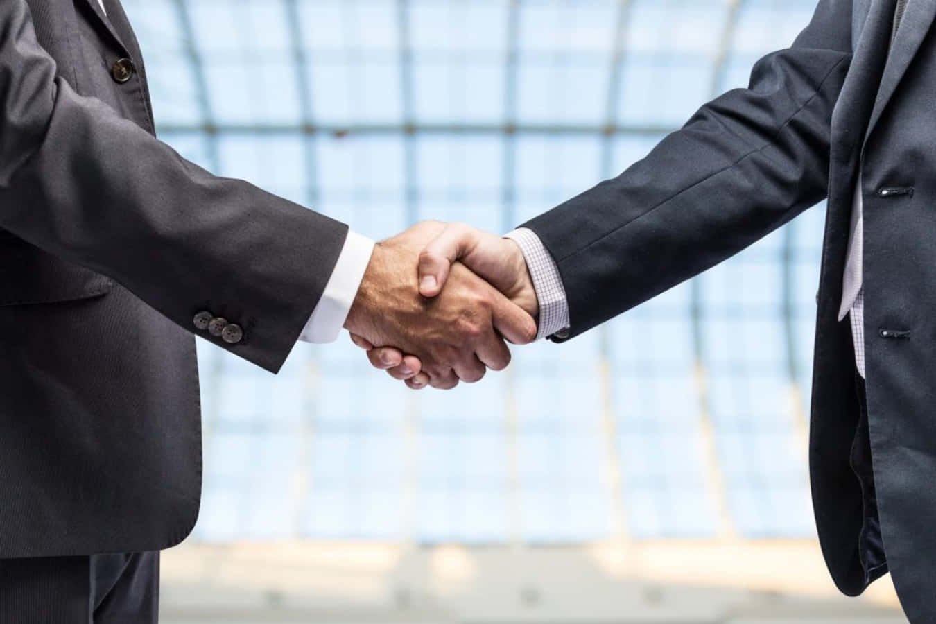 hands 桌布 壁紙 — 兩位身穿深色西裝的商人，在明亮的玻璃面板背景下堅定地握手。