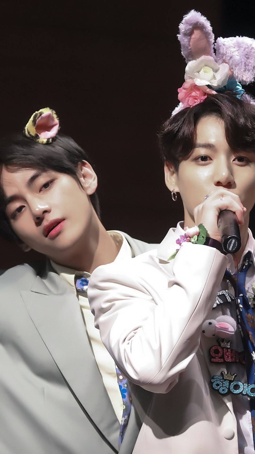 BTS 桌布 壁紙 — 兩位身穿淺色西裝的年輕男子戴著花卉兔耳，其中一人在舞台燈光下對著麥克風唱歌。