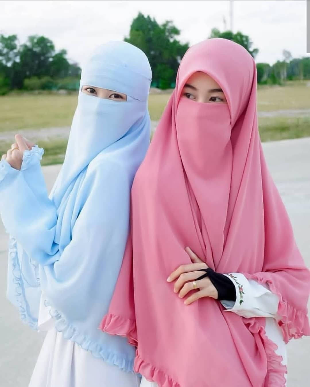 muslim 桌布 壁紙 — 兩位身穿淺藍色和粉紅色尼卡布的女性在戶外擺姿勢，帶著溫柔的微笑和荷葉邊袖子，背景是柔和的綠色。
