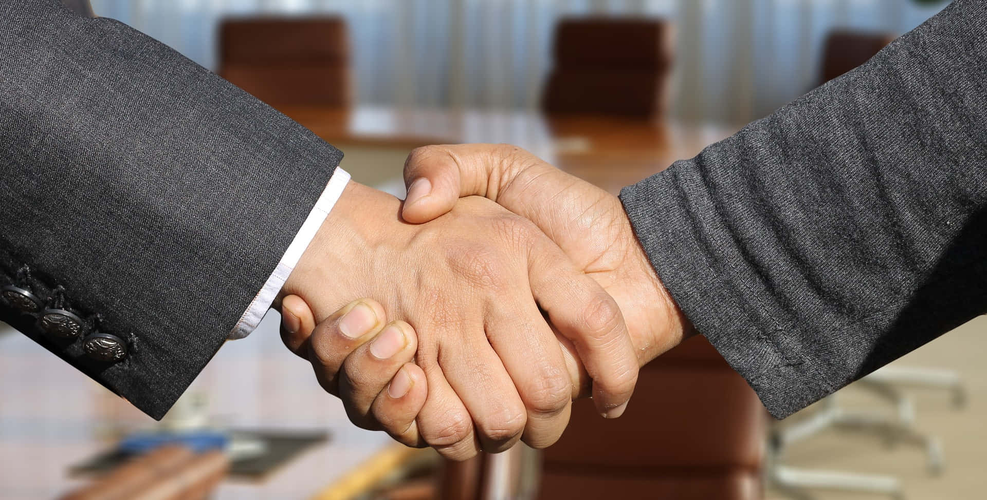 商業 桌布 壁紙 — 兩位身穿灰色西裝的商務人士在現代辦公室中，隔著模糊的會議桌堅定地握手。