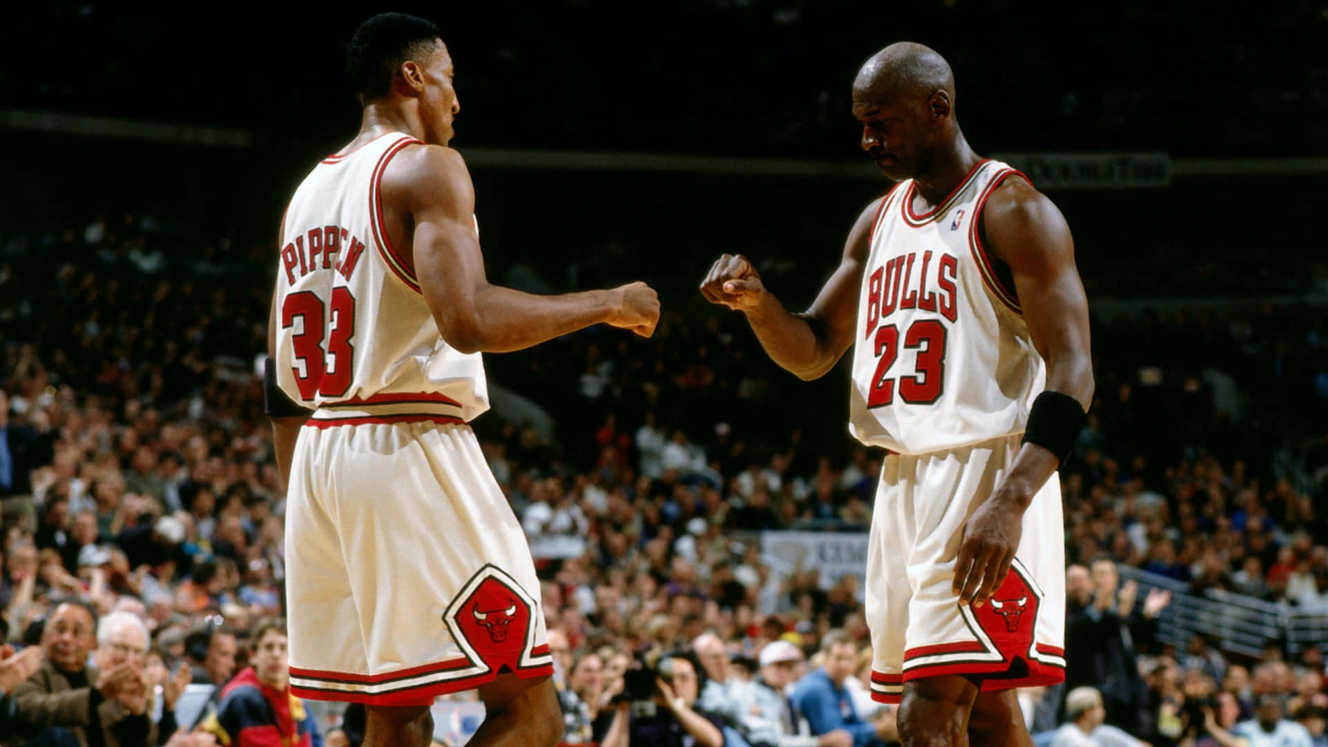 Aesthetic NBA 桌布 壁紙 — 兩位身穿白色球衣的芝加哥公牛籃球運動員在燈光明亮的球場上互相碰拳，身後是歡呼的人群。