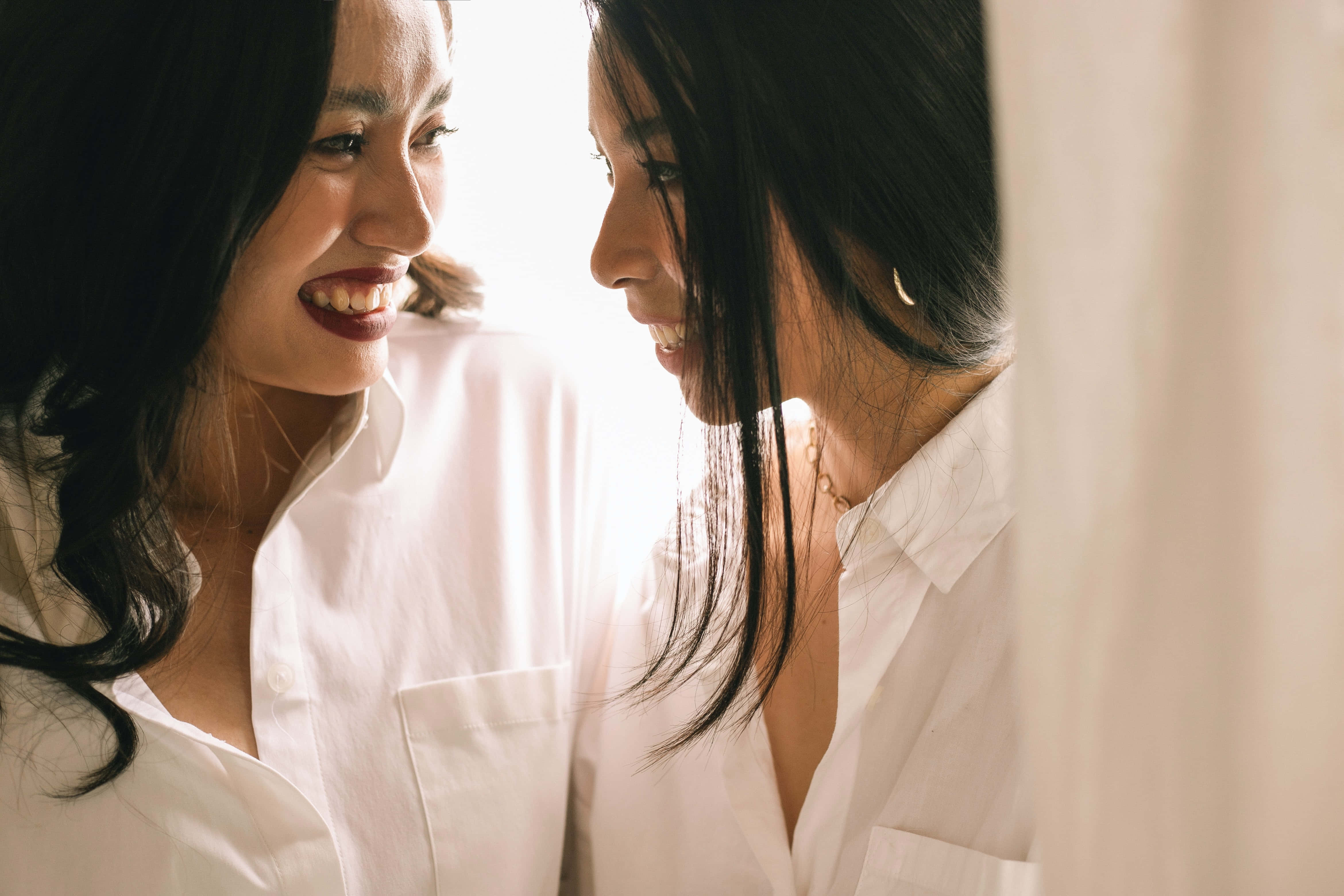 女同志 桌布 壁紙 — 兩位身穿白襯衫的女性在柔和自然光中分享明亮親密時刻，溫暖地微笑相對。