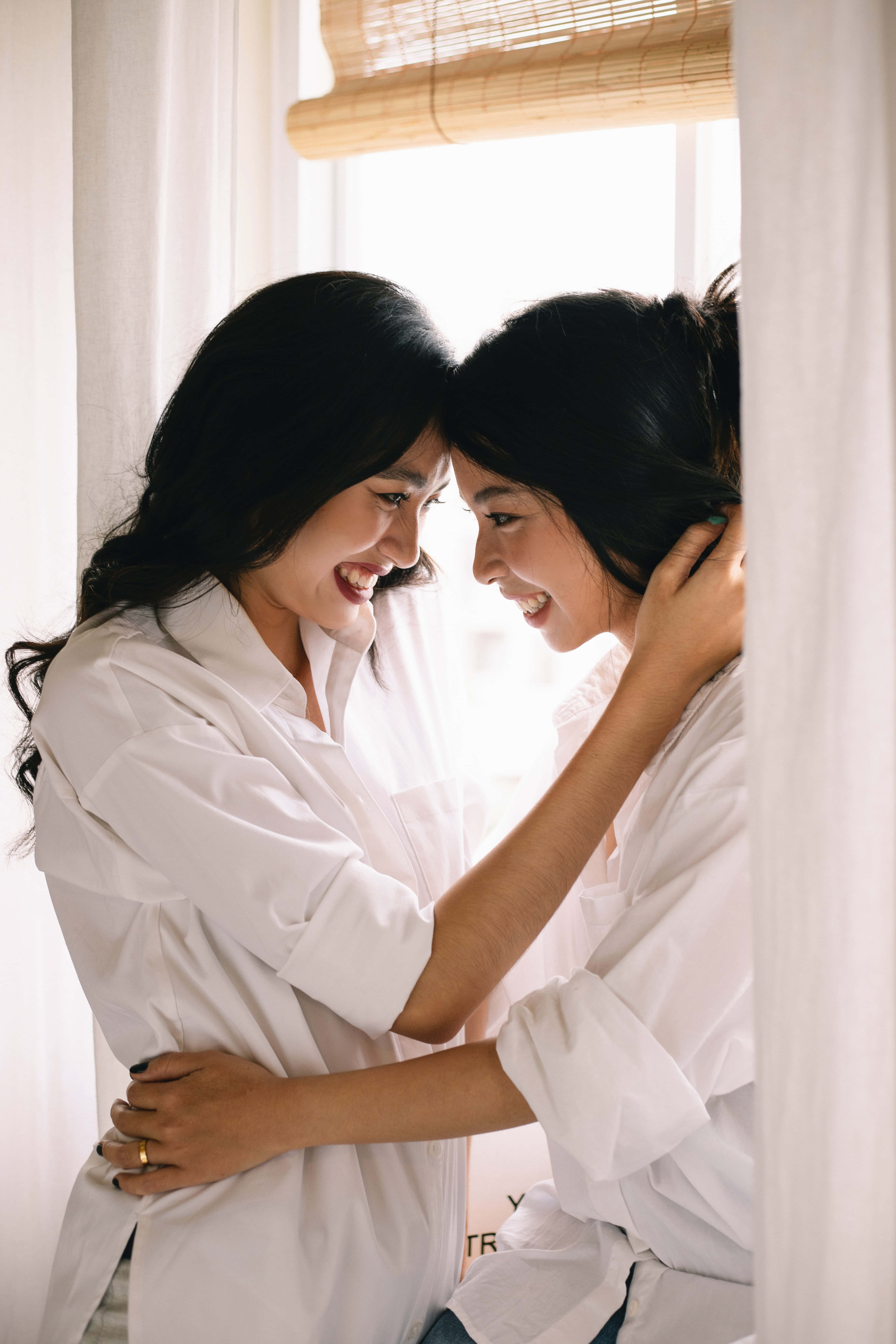 Lesbian Girl 桌布 壁紙 — 兩位身穿白襯衫的女性在竹簾和柔和自然光的窗邊快樂地擁抱。