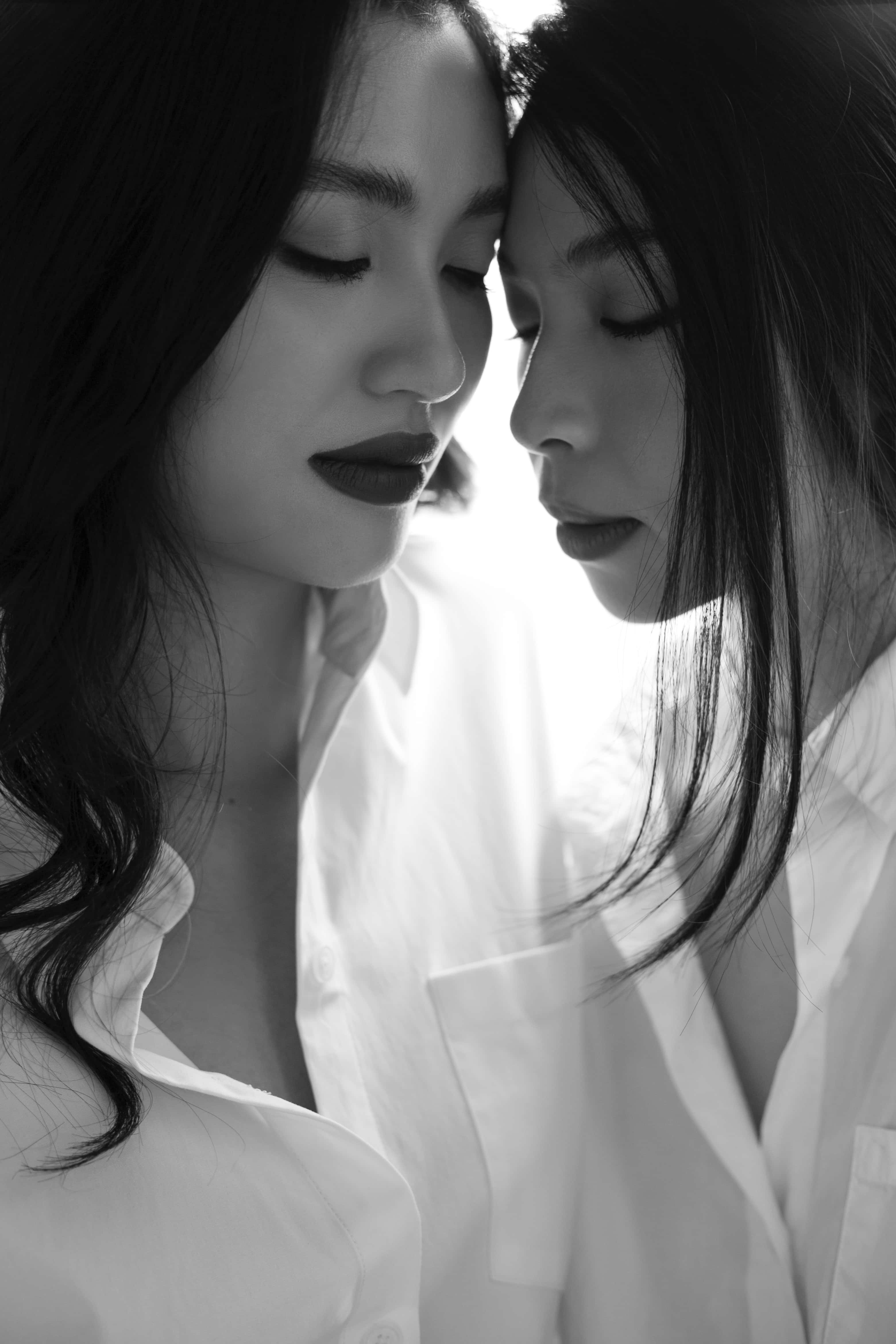 女同志 桌布 壁紙 — 兩位身穿白襯衫的女性額頭相貼，在柔和的黑白肖像照中帶有戲劇性的燈光。