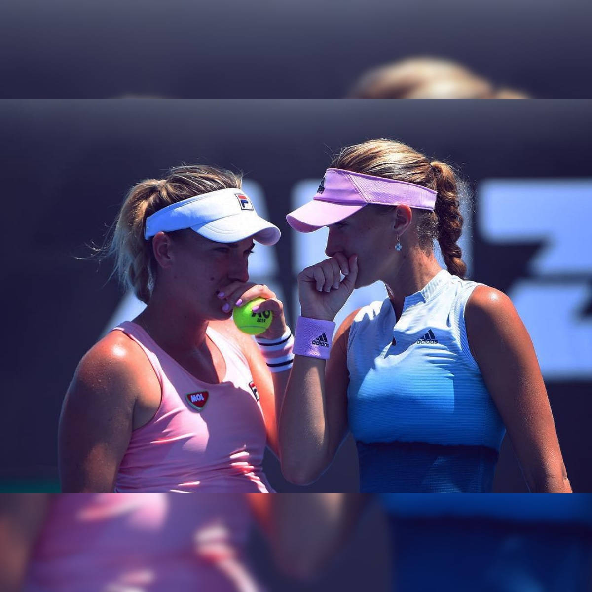 Australian Open 桌布 壁紙 — 兩位身穿粉紅色和藍色服裝的女網球運動員在陽光明媚的球場上共享寧靜時刻，其中一位咬著網球，另一位觸摸嘴唇。