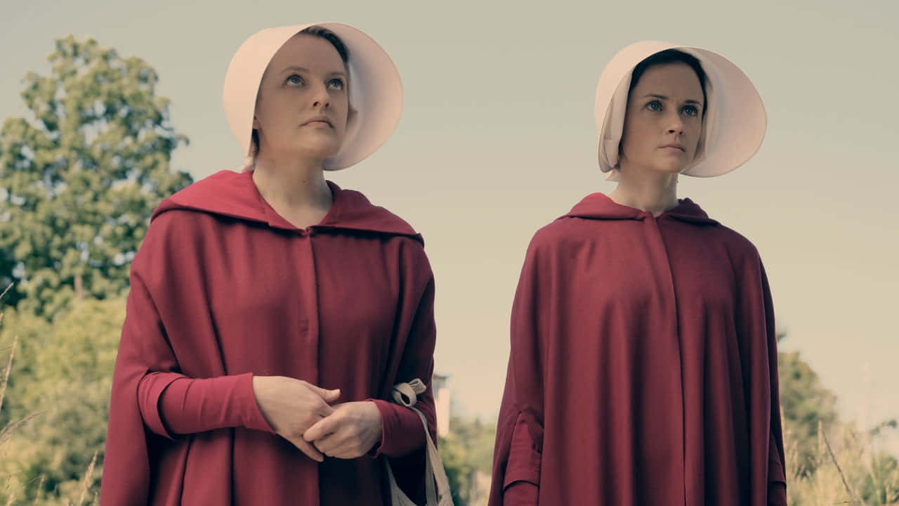 Red Clothing 桌布 壁紙 — 兩位身穿紅色斗篷和白色帽子的女性，莊嚴地站在陽光明媚的田野中，身後有樹木。
