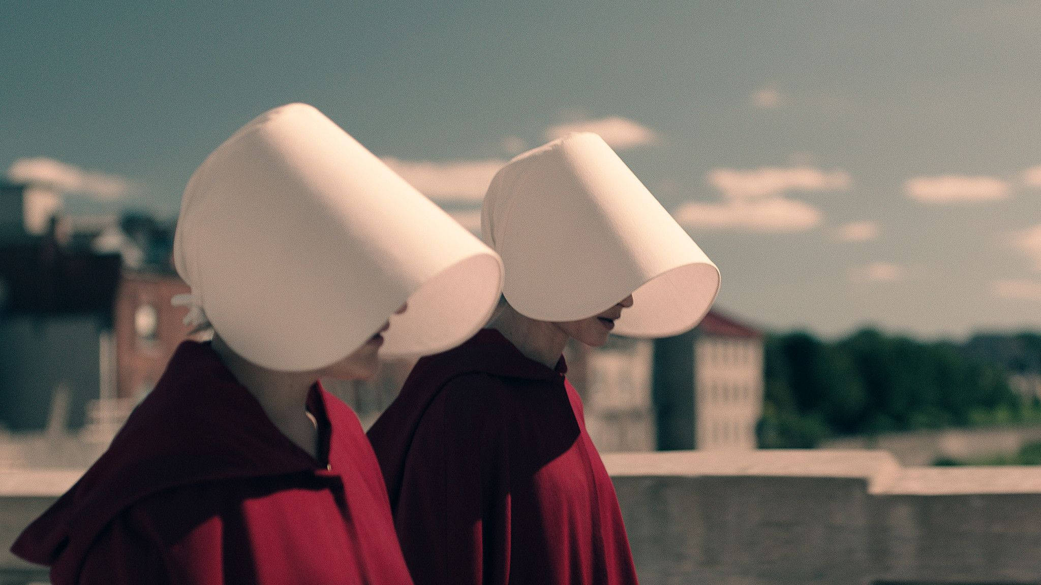 The Handmaid's Tale 桌布 壁紙 — 兩位身穿紅色長袍和白色帽子的女性並肩站在屋頂上，俯瞰著多雲天空下朦朧的城市景觀。