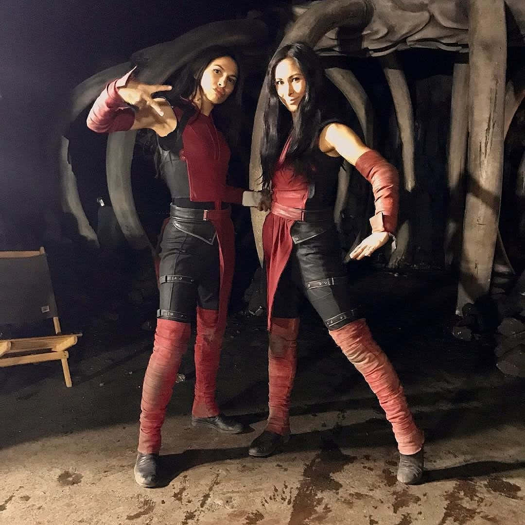 Celebrity 桌布 壁紙 — 兩位身穿紅黑cosplay的女性在燈光昏暗的攝影棚中，自信地站在一個巨大的骷髏道具前擺姿勢。