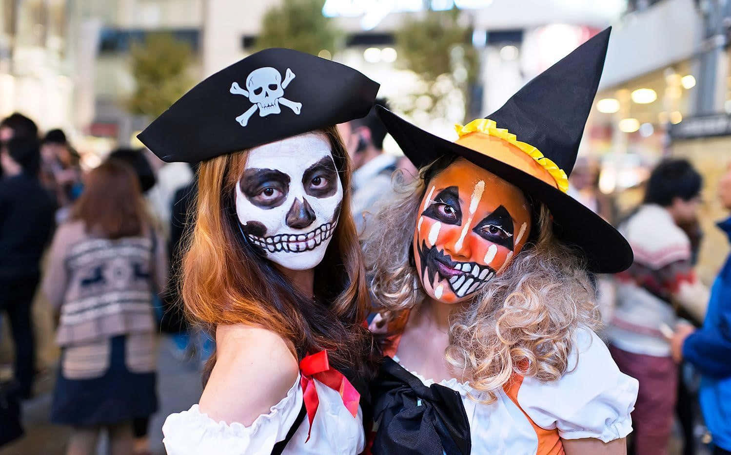 Horror Costumes 桌布 壁紙 — 兩位身穿萬聖節服飾、化上骷髏和女巫妝容的女性，在繁華的都市街道上一起擺姿勢。