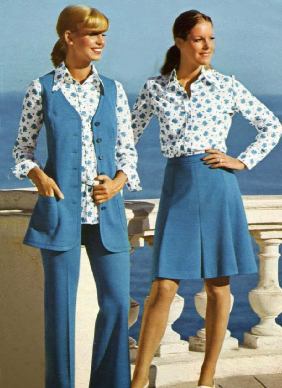 70年代動圖 桌布 壁紙 — 兩位身穿藍色花卉圖案服裝的女性，在晴朗的藍天下，自信地站在可俯瞰海洋的石砌陽台上擺姿勢。